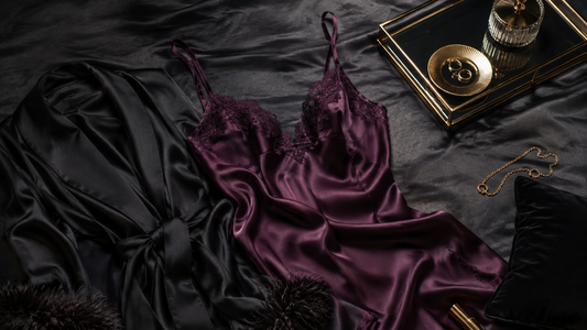 Dark plum satin camisole and black robe arranged with muted gold accessories on charcoal fabric